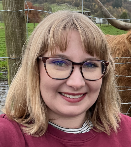 Headshot of a blond woman wearing wood grain toned glasses and a head sweater standing in front of a wire fence